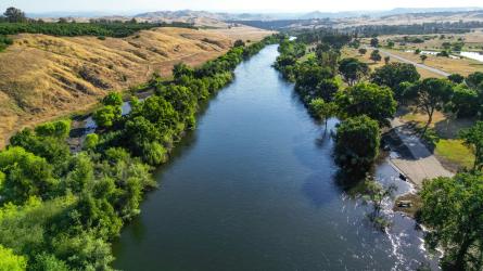 An aerial view shows a river bordered by green trees and dry rolling hills.