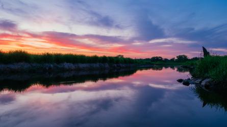 A quiet wetland channel reflects sunset clouds, with grasses along both banks.