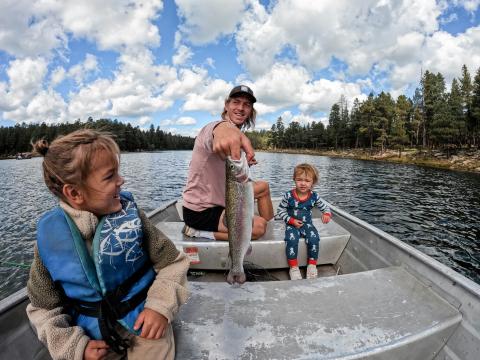 An adult and two children sit in a small boat on a forested lake while the adult holds up a freshly caught fish.
