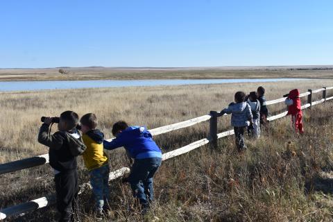 Young students look across wetland habitat from behind a fence, with one student using binoculars.