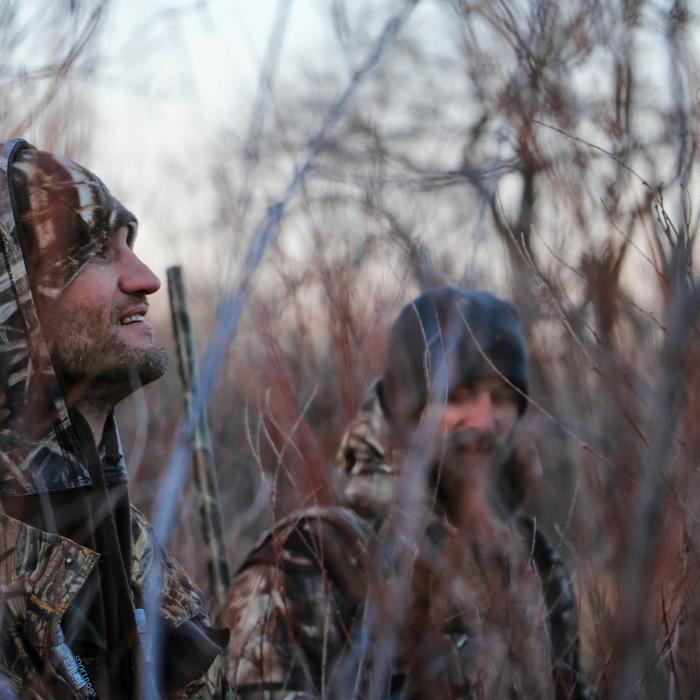 Two hunters in camouflage sit among bare shrubs in a wetland or wooded habitat.