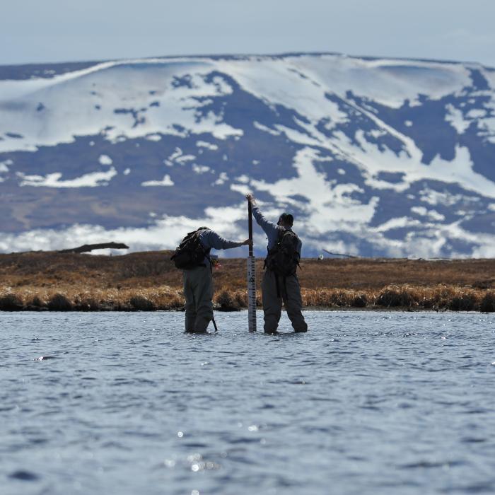 Two field researchers stand in shallow water near a data logger with snow-covered hills in the background.