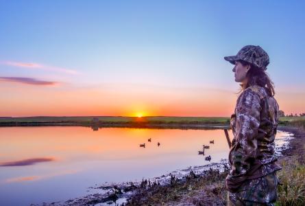A waterfowl hunter stands beside a wetland at sunset with ducks floating on the water.
